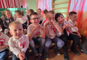 Dzieci siedzą w rzędach i uśmiechają się do zdjęcia; mają przypięte kotyliony i odświętne stroje.