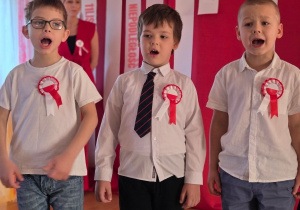 Trzech chłopców śpiewa podczas występu patriotycznego na tle biało-czerwonej dekoracji.
