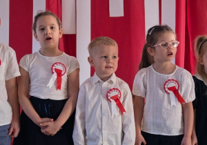 Dzieci stoją w rzędzie i śpiewają piosenkę, przed czerwono-białą dekoracją z hasłami patriotycznymi.