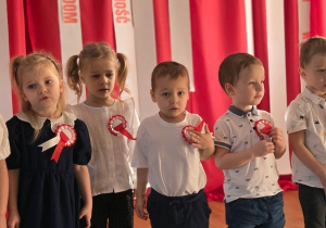 Grupa najmłodszych dzieci stoi w szeregu, każde ma przypięty biało-czerwony kotylion.