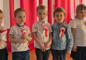 Kilkoro dzieci stoi w szeregu na tle biało-czerwonej dekoracji, każde ma przypięty kotylion.