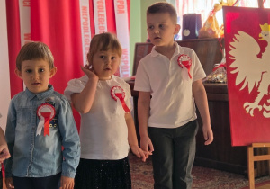 Troje dzieci stoi obok siebie przed patriotyczną dekoracją i tablicą z godłem Polski.