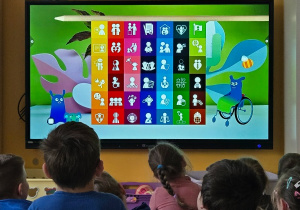 Dzieci siedzą przed ekranem z kolorowymi ikonami przedstawiającymi prawa dziecka.