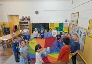 Grupa dzieci stoi wokół kolorowej chusty animacyjnej z balonami.