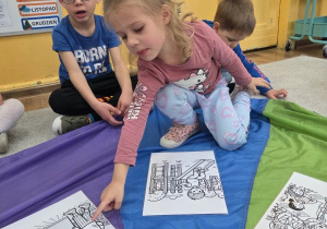 Dziewczynka wskazuje obrazek ułożony na kolorowej chuście, a obok siedzą inne dzieci.