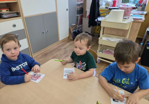 Trzech chłopców siedzi przy stoliku i koloruje małe obrazki.
