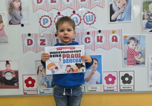 Dziecko stoi przed tablicą z napisem „Dzień Praw Dziecka” i trzyma kartkę z hasłem święta.