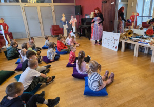 Dzieci siedzą na poduchach i słuchają prowadzącej podczas zajęć muzycznych w teatrze.