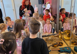 Grupa dzieci ogląda instrumenty muzyczne, a prowadząca pokazuje im trąbkę.