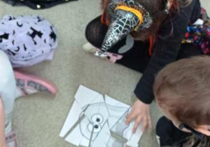 Dzieci układają puzzle z rysunkiem potwora, siedząc w kręgu na podłodze w halloweenowych przebraniach.