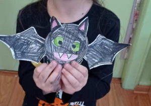 Dziewczynka trzyma w rękach własnoręcznie pokolorowaną papierową ozdobę nietoperza i uśmiecha się.