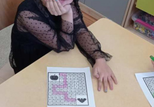 Dziewczynka siedzi przy stoliku i koloruje halloweenową kartę pracy z liczbami.