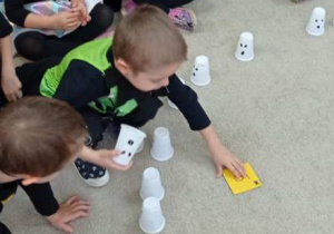 Grupa dzieci bawi się na dywanie kubeczkami z narysowanymi duszkami, uczestnicząc w halloweenowej grze.