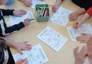 Kilka dzieci siedzi przy stole i koloruje halloweenowe obrazki.