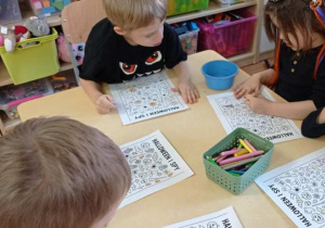 Grupa dzieci przy stoliku wykonuje zadanie „Halloween I Spy”, wyszukując i kolorując obrazki.