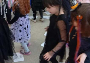 Dzieci maszerują po sali, stawiając stopy na kartkach z halloweenowymi rysunkami ułożonymi na podłodze.
