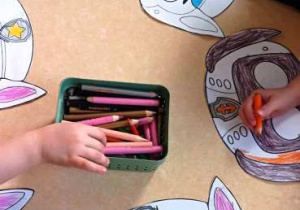 Dzieci kolorują papierowe maski postaci z bajki, siedząc przy stoliku.