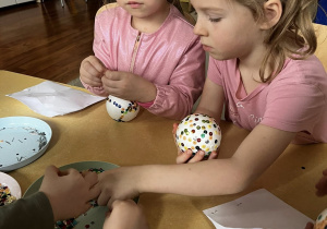 Zdjęcie przedstawiające dzieci robiące bombki ze styropianu i cekinów przy stoliku.