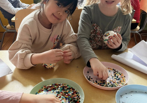 Zdjęcie przedstawiające dzieci robiące bombki ze styropianu i cekinów przy stoliku.