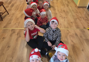Grupa dzieci w czapkach Mikołaja siedzi jedno za drugim na podłodze, uśmiechając się do zdjęcia.