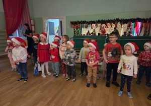 Dzieci w mikołajkowych czapkach stoją w szeregu w sali, przygotowane do występu dla Mikołaja.