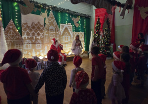 Dzieci w czapkach Mikołaja stoją zwrócone w stronę scenki, gdzie znajduje się Mikołaj i dekoracje świąteczne.