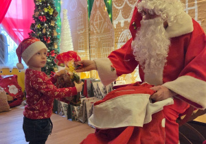 Dziecko w czapce Mikołaja odbiera prezent od Mikołaja siedzącego przy świątecznej scenografii.