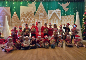 Grupa dzieci w świątecznych strojach i czapkach Mikołaja pozuje z prezentami obok Mikołaja na tle zimowej dekoracji.