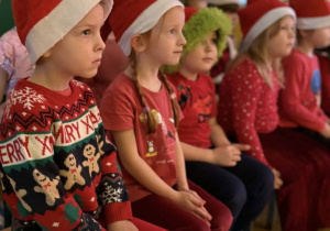 Dzieci w czapkach Mikołaja siedzą na ławeczkach i słuchają św. Mikołaja.