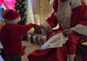 Dziecko w czapce Mikołaja podaje rękę Świętemu Mikołajowi podczas wręczania prezentu.