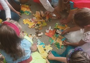 Grupa przedszkolaków siedzi na dywanie i wspólnie układa duże, kolorowe puzzle z postaciami z bajek. Dzieci mają na sobie stroje księżniczek i bajkowych bohaterów.