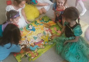 Dzieci siedzą w kręgu wokół ułożonych puzzli przedstawiających bohaterów bajki o Królewnie Śnieżce i krasnoludkach. Przedszkolaki przyglądają się gotowemu obrazkowi i rozmawiają.