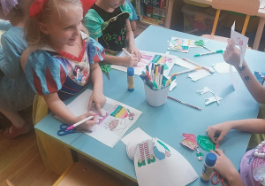 Dzieci siedzą przy stoliku i wykonują prace plastyczne. Kolorują, wycinają i przyklejają papierowe elementy. Na stole znajdują się kredki, klej i nożyczki.