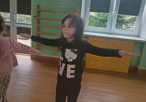 Dziewczynka stoi na sali gimnastycznej z wyciągniętymi rękami i wykonuje ćwiczenie równoważne z woreczkiem na głowie. W tle widoczne są inne dzieci.