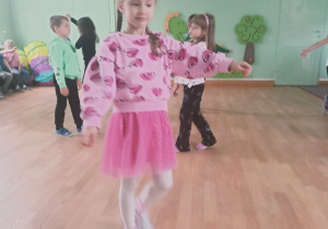 Dziewczynka idzie po sali gimnastycznej z rozłożonymi rękami. Podczas ćwiczenia woreczek gimnastyczny spadł jej z głowy. W tle ćwiczą inne dzieci.