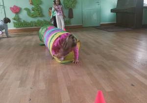 Dziecko czołga się przez kolorowy tunel w trakcie toru przeszkód. Na podłodze stoi pachołek wyznaczający trasę.