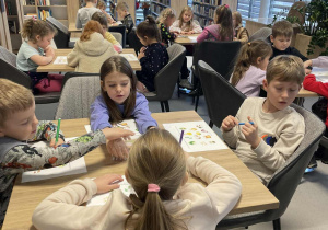 Dzieci siedzą przy stolikach i pracują plastycznie w bibliotece.