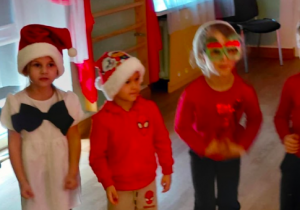 Dzieci w czerwonych ubraniach i czapkach Mikołaja stoją w sali i przygotowują się do występu.