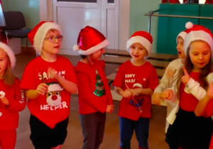 Grupa dzieci w czapkach Mikołaja śpiewa i gestykuluje podczas spotkania świątecznego.