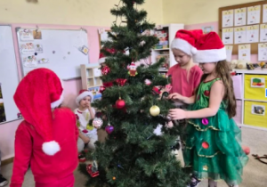 Dzieci w czapkach świątecznych wspólnie ubierają choinkę w sali przedszkolnej.