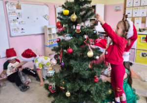 Dziewczynka w czerwonym stroju wiesza ozdobę na choince.