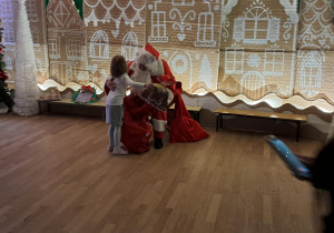 Dziewczynka podchodzi do Mikołaja, który wręcza jej prezent na tle świątecznej dekoracji.