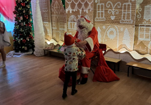 Mikołaj siedzi na krześle i rozmawia z dzieckiem, obok stoi choinka.