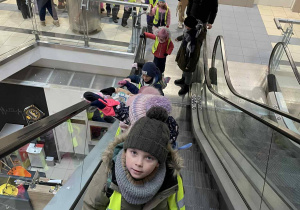 Grupa dzieci w kamizelkach odblaskowych jedzie ruchomymi schodami w budynku publicznym.