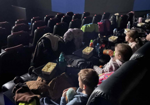 Dzieci siedzą w sali kinowej, jedzą popcorn i oglądają film.