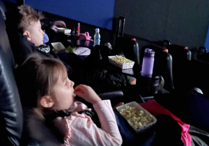 Dzieci w kinie siedzą w fotelach z przekąskami podczas seansu.