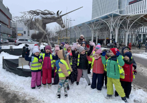 Grupa dzieci w kamizelkach odblaskowych stoi na śniegu przy rzeźbie jednorożca w centrum miasta.