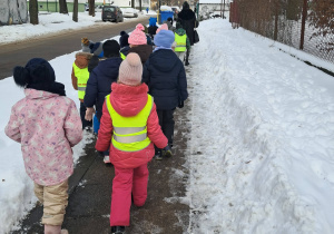 Grupa dzieci w kamizelkach odblaskowych idzie chodnikiem w zimowej scenerii miasta.