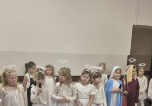 Dzieci przebrane za anioły i postacie z jasełek stoją przed publicznością podczas występu.