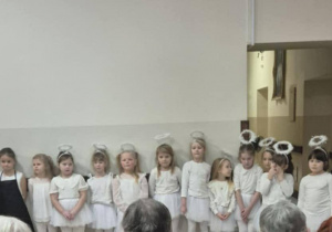 Dzieci w białych strojach aniołów stoją w szeregu, przed nimi siedzą seniorzy.